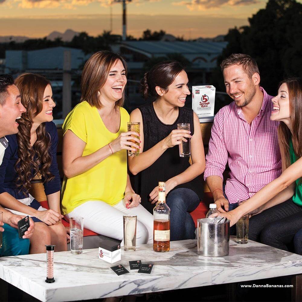 Six friends laugh and enjoy drinks on a patio, transforming their casual sunset gathering into an impromptu game night. A woman reaches for a card from the ice bucket while bottles and glasses fill the table, setting the stage for Danna Bananas' 5 Second Rule Uncensored Game.