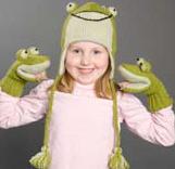 A smiling child wears the Froggy Pilot Hat by Danna Bananas, a green frog-themed hand-knit wool accessory with dangling tassels and matching mittens. This adorable fair trade certified set is set against a neutral gray background.