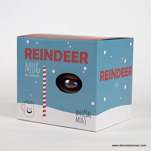 The packaging box, in blue and white, highlights the Reindeer Heat Change Mug from Danna Bananas, making it an ideal Secret Santa gift. It includes red text and a small window that displays part of the mug inside, decorated with snowflakes and candy cane motifs.