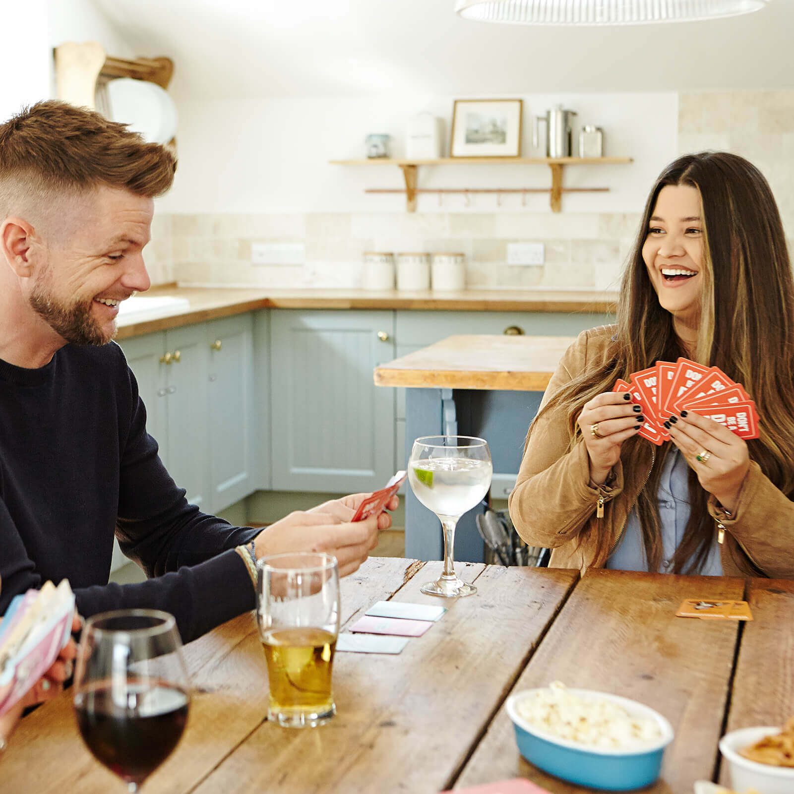 Two friends laughing and playing "Don't Be A Dik Dik" card game at a kitchen table with drinks and snacks.