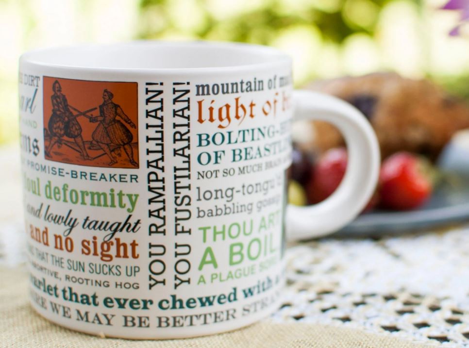A Danna Bananas Shakespeare Insults Mug, featuring colorful Shakespearian insults such as "you rampallian!" and "light of brain," sits atop a lace tablecloth, with blurred fruits in the background. It's the perfect gift box addition for literary enthusiasts.