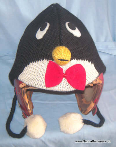 The Penguin Pilot Hat - Kids by Danna Bananas features big eyes, a yellow beak, red bow tie, and ear flaps with black braided ties ending in white pom-poms. Made from New Zealand wool with a light blue background, it's perfect for any adventurous child.