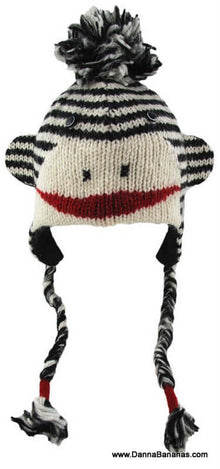 The Sock Monkey Pilot Hat by Danna Bananas is a playful kids' hat made of New Zealand wool, featuring black and white stripes, a red mouth, button eyes, and pom-poms on the top and earflap ends.