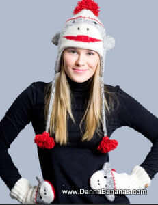 A person wears a Danna Bananas Sock Monkey Pilot Hat - Grey Adult with earflaps and matching mittens, crafted from New Zealand wool in red, white, and gray with pom-poms, supporting fair trade practices, against a plain background.