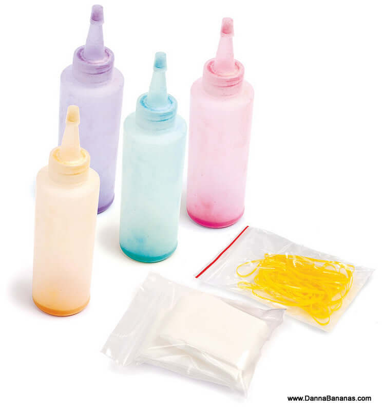 Four upright squeeze bottles in purple, blue, pink, and peach from the Outset Media Tie Dye Kit are displayed. In front of them, two plastic bags are placed: one containing yellow rubber bands and another with a white powdery substance ideal for non-toxic tie dye projects.