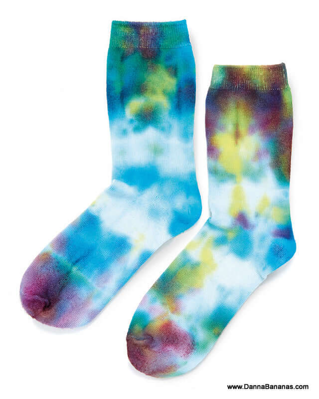 A pair of tie-dye socks featuring a lively blend of blue, yellow, green, and purple patterns crafted using the Outset Media Tie Dye Kit. Made with non-toxic materials, the vivid and artistic design pops against a plain white backdrop. Ideal for individuals looking for hypo-allergenic footwear options.