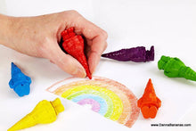 A hand uses a red Bavarian Gnome Crayon from Danna Bananas to color a rainbow on white paper. Six other vibrant crayons, crafted from non-toxic wax in shades of blue, yellow, orange, green, purple, and teal, are arranged around the drawing.