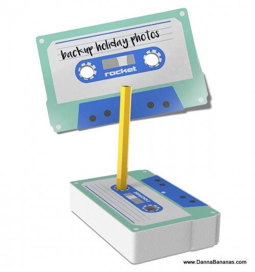 Introducing the Cas-Set Sticky Notes Set by Danna Bananas—a note holder shaped like a cassette that comes with retro-inspired sticky notes. It features a yellow pencil in the middle, reminiscent of a tape reel, and bears a label that reads "backup holiday photos." The base replicates the iconic design of a classic cassette tape, making it perfect for adding a nostalgic touch to your desk.