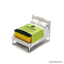 The Clean Dreams Kitchen Sponge Holder by Danna Bananas showcases a white miniature bed design with a yellow sponge as the mattress, complete with a green abrasive side. Wrapped in a paper label, this playful creation resembles a bedspread and cleverly transforms into a distinctive kitchen sponge holder.
