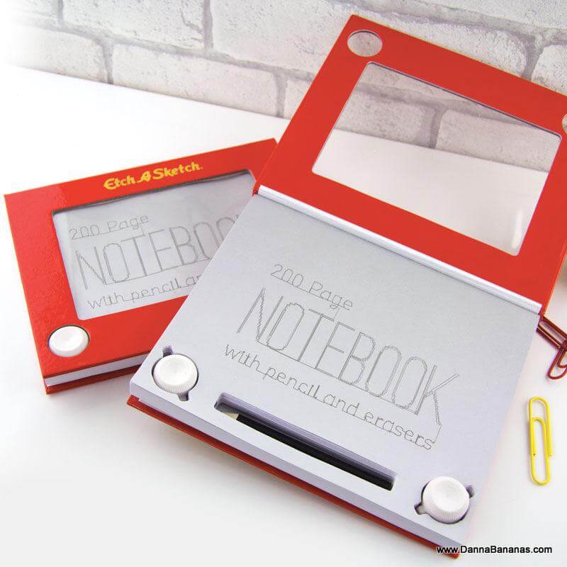 Two Etch A Sketch NoteBooks by Danna Bananas are open. The notebook on the left displays "200 Page Notebook" in a font akin to an Etch A Sketch, accompanied by a pencil and erasers. The one on the right has its cover partially open with two paper clips next to it.