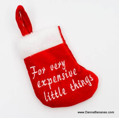 The "Christmas Stocking for Very Expensive Things" by Danna Bananas features a red stocking with a white cuff and white embroidered text that reads, "For very expensive little things." This slightly angled stocking comes with a loop at the top, perfect for hanging by the fireplace on Christmas morning. For more details, visit www.DannaBananas.com, displayed in the bottom right corner.