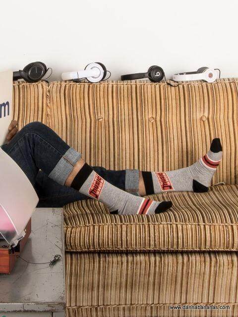 On a striped sofa, a music-loving dad shows off his Giant Music Snob Men's Crew Socks by Danna Bananas beneath rolled-up jeans. Nearby, headphones eagerly await the next playlist session.