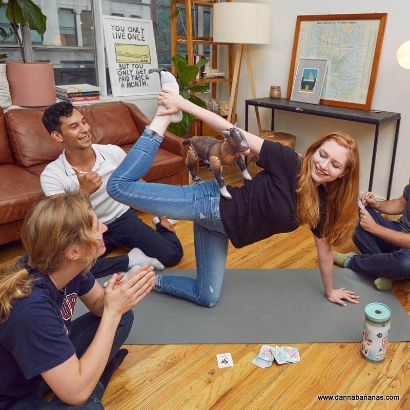 A group of people is enjoying the Goat Yoga Party Game by Danna Bananas in the living room. One person balances gracefully, reminiscent of yoga poses, with a small toy goat perched on their raised leg. Nearby, friends sit smiling and clapping. The room features plants, a camera, and a motivational sign on the wall.