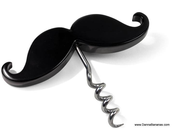 A Handlebar Corkscrew & Bottle Opener from Danna Bananas, featuring a black mustache-shaped design with a shiny silver spiral, is laying on a white surface. It combines functionality with a playful, quirky look, reminiscent of Movember Canada.