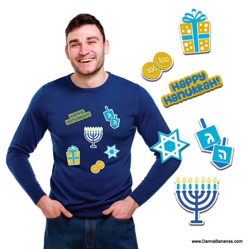 A cheerful man dons the Hanukkah Instant Ugly Sweater Kit by Danna Bananas, featuring vibrant Hanukkah-themed graphics including a menorah, Star of David, dreidels, coins, and a gift. The text "Happy Hanukkah!" is highlighted with festive embroidered patches for an extra holiday flair.