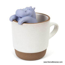 A ceramic mug with a matte beige finish and darker brown base features the playful Danna Bananas Hippo Tea Infuser, which hangs over the rim, perfect for steeping loose leaf tea.