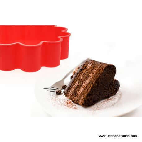 A slice of chocolate cake with cocoa powder dusting rests on a white plate beside a fork, while the vibrant red I Heart Cake Mold by Danna Bananas is ready in the background for your next baking adventure.