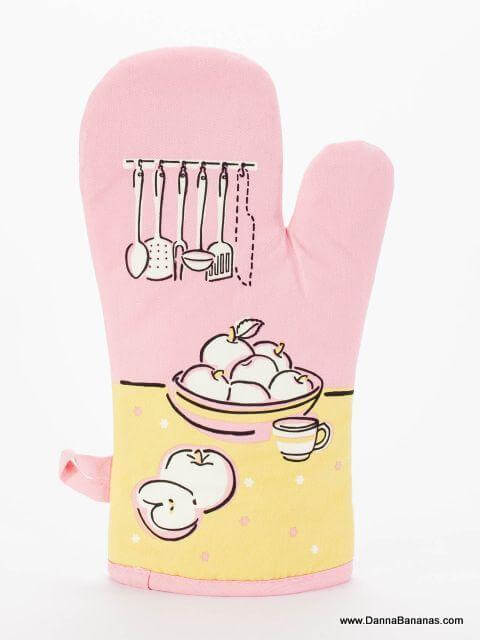 The "I've Got A Knife Oven Mitt" by Danna Bananas is a charming pink kitchen accessory with quilted insulation. It features a fruit bowl, sliced fruit, and cup on a sunny yellow background, with elegant kitchen utensils hanging above the bowl.