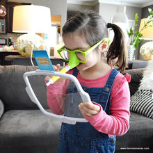 A young girl wearing oversized green glasses draws on a clear clipboard with a pen featuring the signature yellow elephant trunk, similar to the Danna Bananas Pencil Nose Game. Seated in the living room, she brings family game fun to life amidst sofas and cushions in the background.