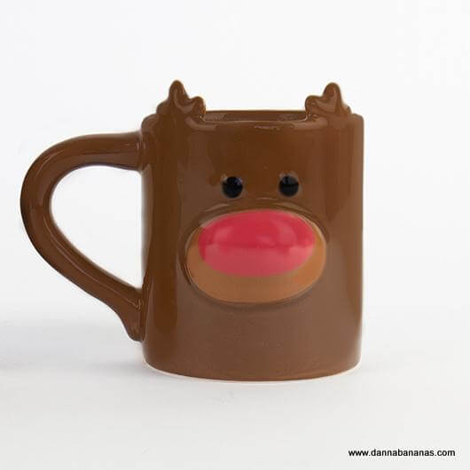 The delightful Reindeer Heat Change Mug by Danna Bananas, ideal for a Secret Santa gift, features raised antler-shaped details on the rim and a cheerful face with black eyes and a bright red nose.