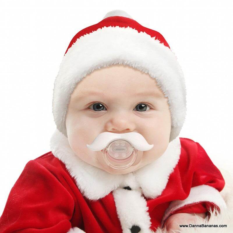 A baby dressed as Santa in a red outfit with fluffy white trim and hat sports the Santafier Mustache Pacifier by Danna Bananas, adding a humorous touch to enchanting holiday photos. The baby's bright eyes and expression perfectly capture the festive, playful theme.