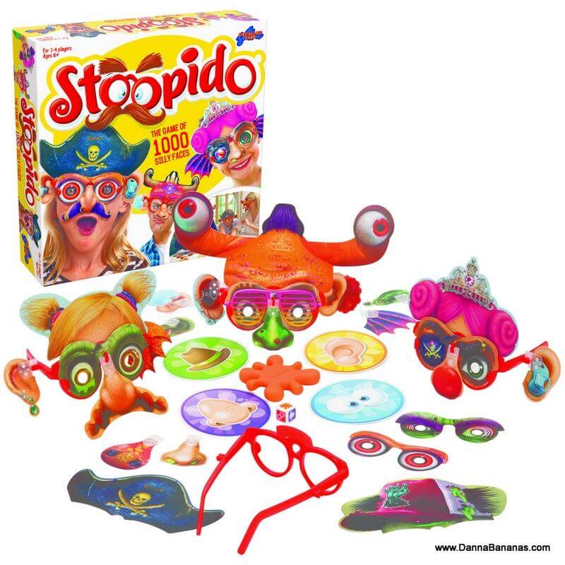 The Stoopido Game by Danna Bananas, a wonderful family game and ideal Christmas gift, includes a vibrant set of quirky face pieces such as glasses, noses, ears, and hats. The box in the background features two individuals adorned with mixed and amusing facial accessories in this entertaining game of silly faces.