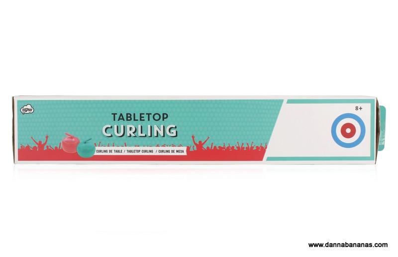 Box for the Danna Bananas Tabletop Curling Game, showcasing a blue and red design with curling graphics on the curling sheet. The text specifies that it is suitable for ages 8 and up. The box displays the brand logo and website URL.