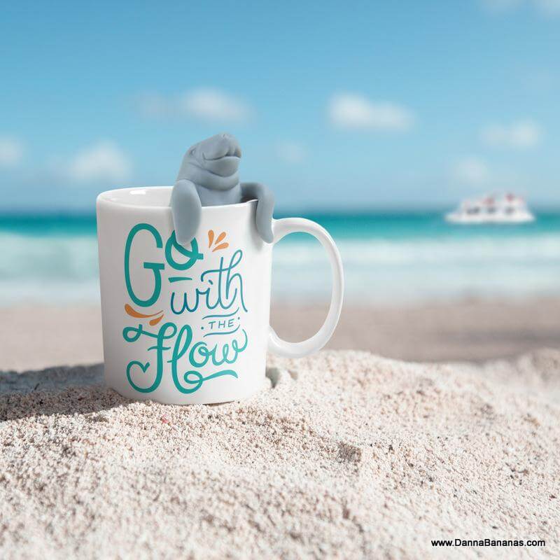 A "Go With The Flow Two For Tea Manatea Mug" by Danna Bananas sits on a sandy beach with a playful tiny shark figure emerging from it, against a backdrop of clear blue sky meeting a turquoise ocean and a distant small ship sailing.