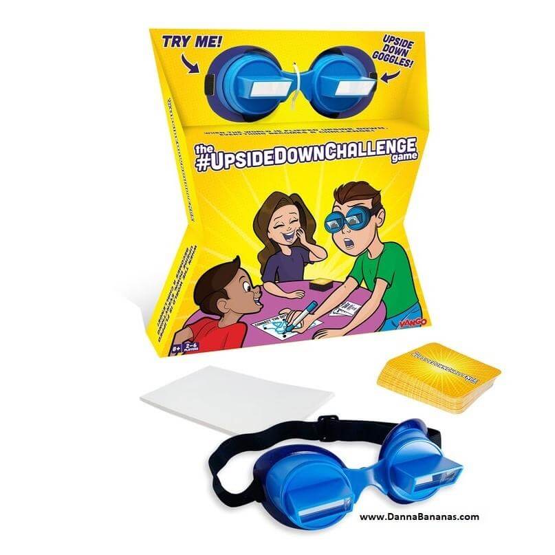 The image showcases the "The UpsideDownChallenge Game" box by Danna Bananas, highlighting a cartoon of people humorously tackling tasks. Next to the box are a pair of blue upside-down goggles, a white challenge card, and a stack of yellow cards—an ideal addition for memorable board game nights. #UpsideDownChallenge