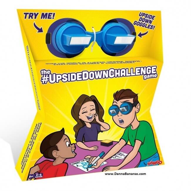 The UpsideDownChallenge Game" by Danna Bananas comes in a yellow box with vibrant illustrations and text. On top, there's a pair of blue upside down goggles. The packaging showcases three people having fun — one wearing the goggles and drawing while the others offer lively reactions, making it an ideal choice for board-game nights.