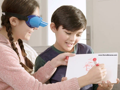 A smiling girl wearing upside down goggles and holding a protruding plastic toy assists a boy who is writing on a whiteboard with a red marker. Embracing The UpsideDownChallenge Game by Danna Bananas, they both appear to be enjoying themselves in the bright room.